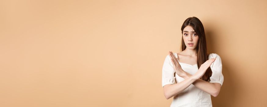 Girl says no. Skeptical young woman disagree or prohibit action, showing cross stop gesture, make nope sign, disapprove bad idea, standing on beige background.