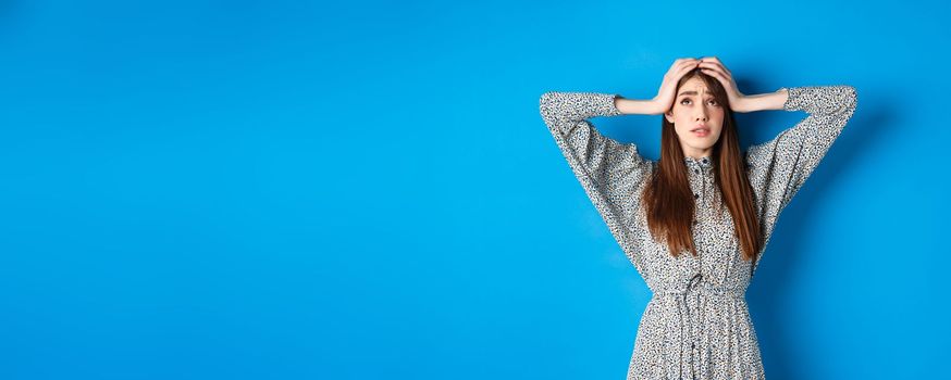 Troubled young woman with long hair, holding hands on head in panic, looking up nervous, having problem, standing on blue background.