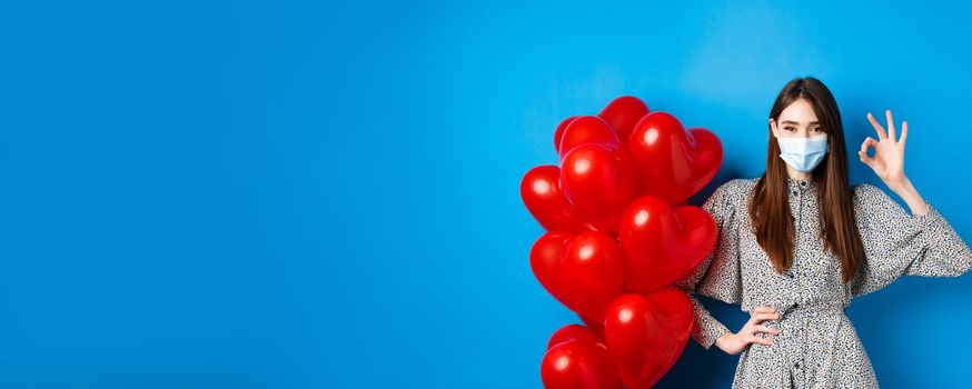 Covid-19, Valentines day and pandemic concept. Beautiful young woman in face mask and dress, showing OK sign in approval, standing near romantic red hearts balloons, blue background.