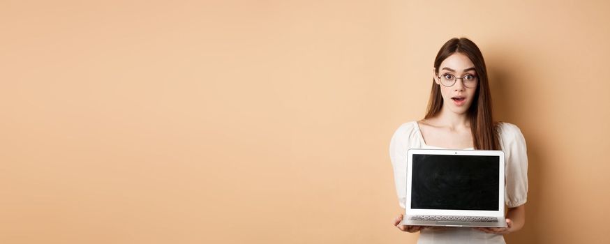 Surprised young woman in glasses showing laptop screen with amazed face, standing on beige background.