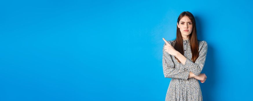 Disappointed frowning girl pointing left and looking displeased, condemn something bad, complaining at banner, standing against blue background in dress.