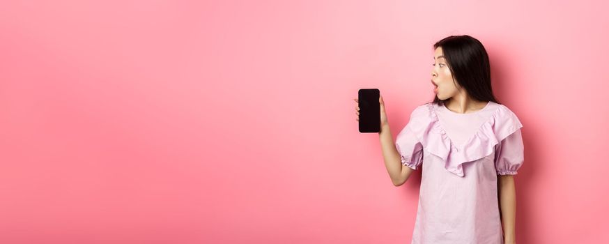 Wow look here. Excited asian girl look at smartphone screen with amazed face, checking out online offer, standing against pink background.