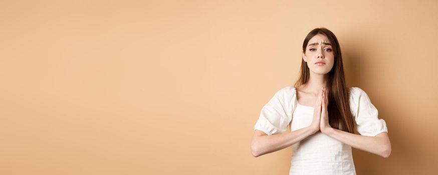 Please help. Desperate young woman begging you, holding hands in pleading gesture and looking sad at camera, asking for favour, beige background.