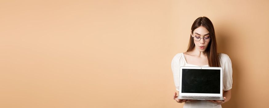 Excited girl in glasses show laptop screen, demonstrate online offer on computer, standing against beige background.