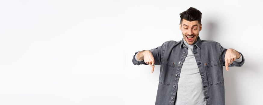 Excited european man looking and pointing fingers down, looking happy at advertisement, standing in denim jacket on white background.