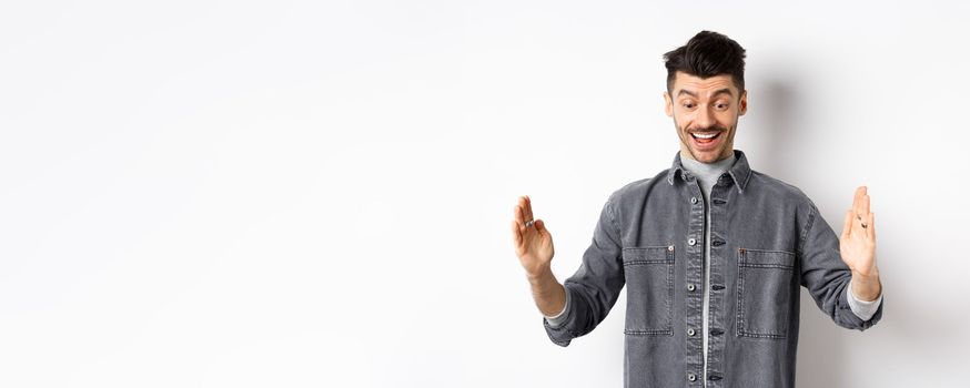 Excited caucasian man showing big size object, shaping large thing and smiling amazed, standing on white background.