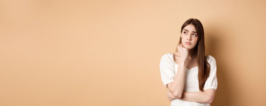 Sad young woman looking pensive at upper left corner, feeling upset or distressed, standing lonely on beige background.