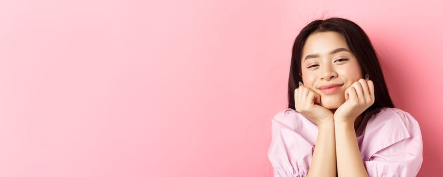Close-up portrait of dreamy and romantic asian girl, lean face on hands and smiling, looking with admiration and happiness, standing against pink background.