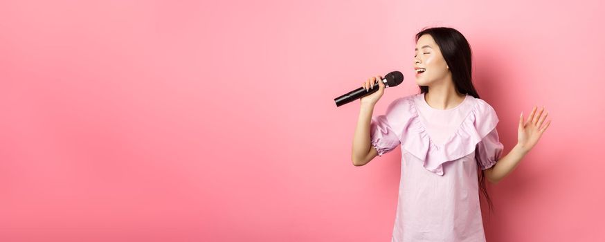 Beautiful asian girl perform song, singing in microphone and smiling romantic, standing in dress against pink background.