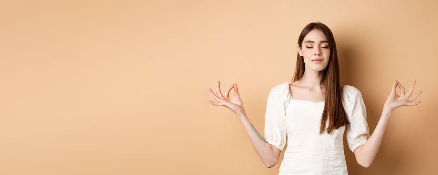 Relaxed young woman meditating, holding hands in yoga zen pose and close eyes, feel peace and smiling, standing on beige background.