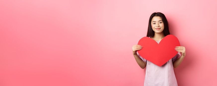 Valentines day concept. Cute teenage asian girl showing big red heart card, falling in love, going on romantic date in dress, smiling tender at camera, pink background.