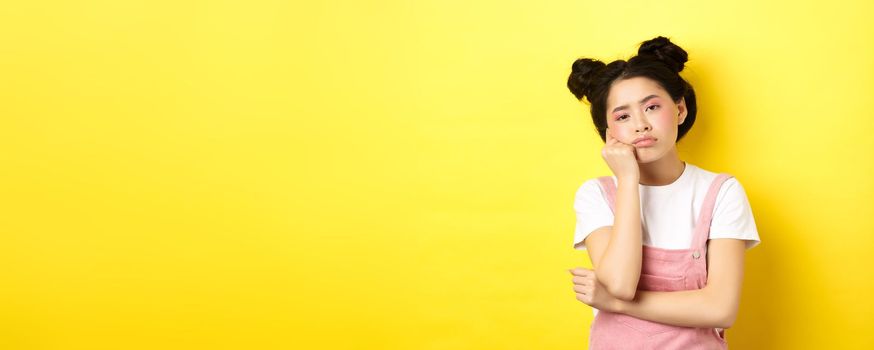 Sad and bored teen asian girl standing alone, looking at camera indifferent, staring with boredom, yellow background.