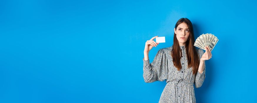 Shopping. Thoughtful girl looking up and biting lip, counting in mind or thinking, holding plastic credit card with dollar bills, standing on blue background.