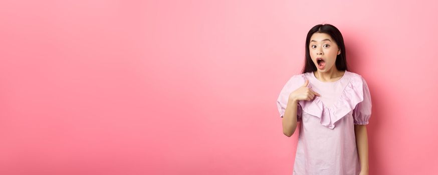 Surprised asian girl gasping wondered, pointing at herself, being chosen or picked, standing in dress against pink background.
