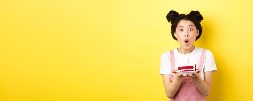 Holidays and celebration. Excited asian birthday girl blowing candle on party cake, making wish on b-day, standing against yellow background.