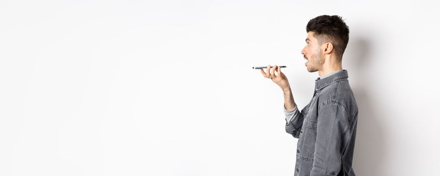 Profile of young man speaking at smartphone, translate with voice-translator app, talking into mobile phone speakerphone, standing on white background.