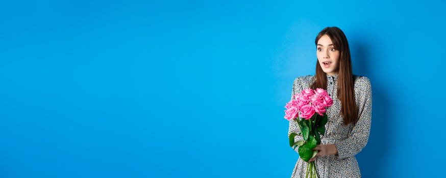 Valentines day concept. Surprised happy girl looking aside with amazement, receiving bouquet of romantic flowers, holding pink roses, standing in dress on blue background.