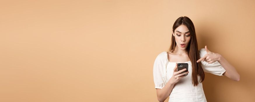 Exciting news on phone. Surprised cute woman reading online promo on screen, pointing at smartphone and say wow amazed, standing on beige background.