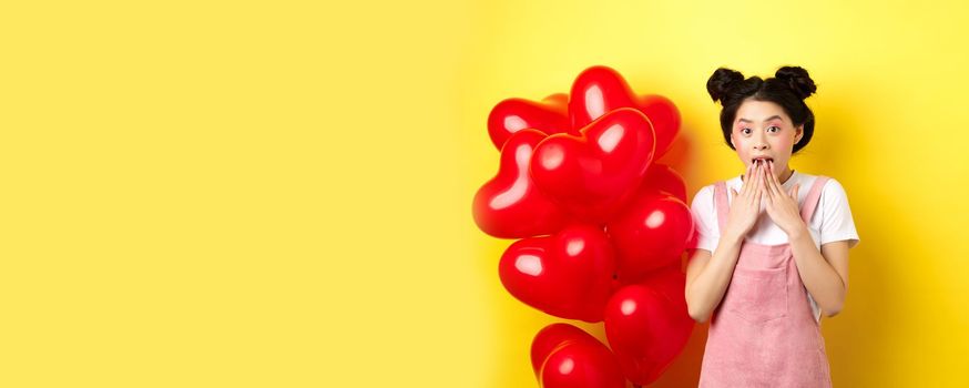 Surprised korean girl gasping and saying wow, looking at special promo offer on Valentines day, standing near big red hearts balloons and yellow background.