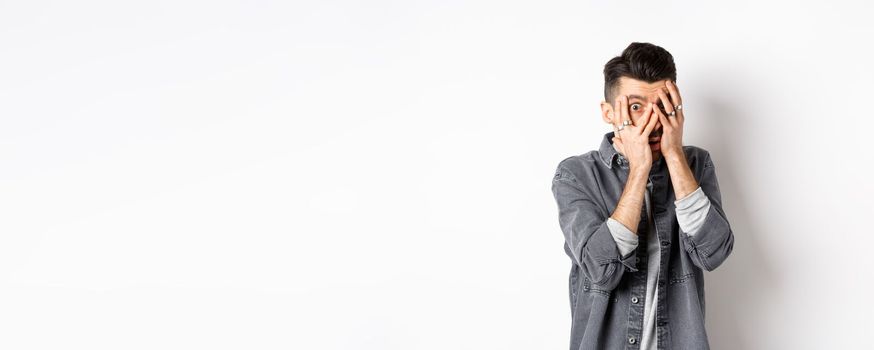 Scared young man peeking through fingers with nervous face, staring at something scary, standing on white background.