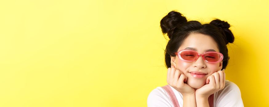 Summer fashion concept. Dreamy asian girl with romantic face expression, daydreaming or imaging something beautiful with closed eyes and happy smile, wearing sunglasses.