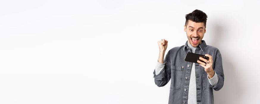 Happy man shouting yes, winning video game on mobile phone and triumphing, achieve online goal, standing on white background.