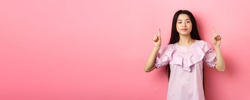 Smiling teen girl asian pointing fingers up at empty space, advertising on pink background.