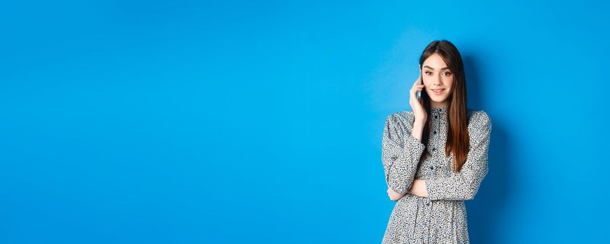 Stylish young woman talking on phone, making takeout order via smartphone, standing in dress against blue background.
