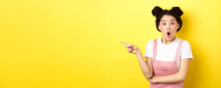Surprised asian teen girl looking impressed, say wow and pointing finger left, standing with glamour makeup and summer clothes on yellow background.