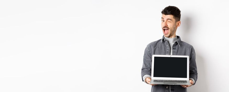 Cheerful guy winking and showing empty smartphone screen, offer you advertisement, standing on white background.
