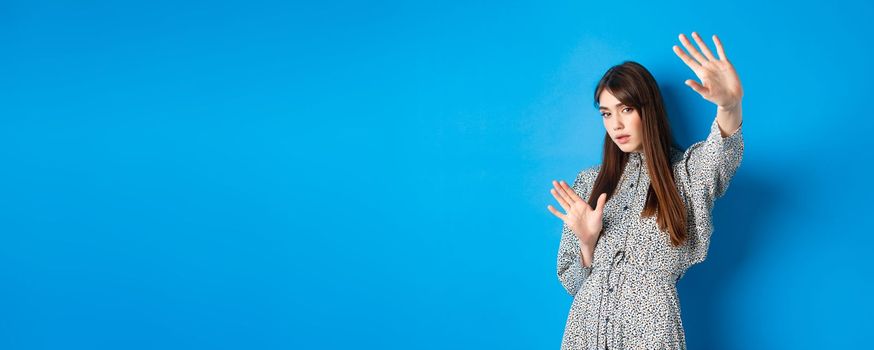 Image of young woman raising hands up defensive, covering herself from camera fleshlights, standing on blue background. Copy space