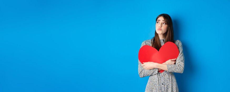 Valentines day. Sad heartbroken girl sobbing, looking aside at empty space and crying upset, hugging big red heart, feeling lonely without lover, standing alone on blue background.