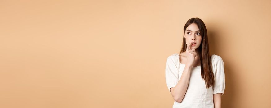 Nervous young girl biting fingernail and looking aside excited, making choice, thinking about something, standing on beige background.