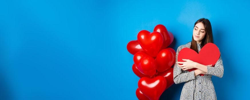 Valentines day. Romantic pretty woman in dress hugging big red heart cutout and looking dreamy, thinking of love, standing on blue background.