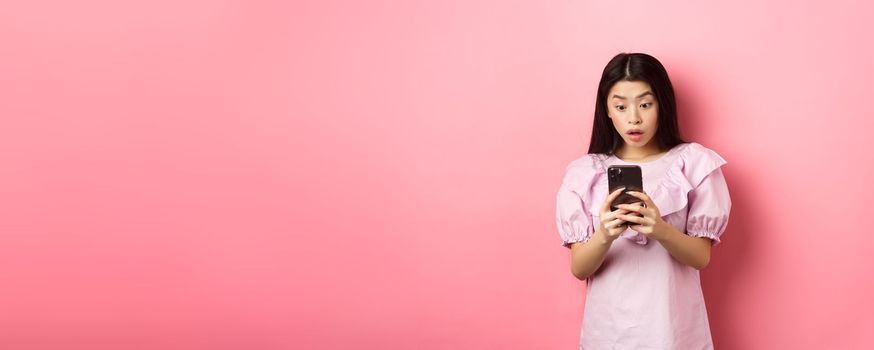 Online shopping. Asian teen girl look surprised at mobile phone screen, reading message with excited expression, standing against pink background.