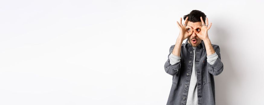 Funny guy look through hand binoculars, checking out awesome promo, say wow and stare at logo on left with excited face, standing on white background.