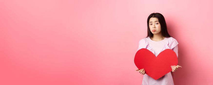 Valentines concept. Single teenage asian girl wants to fall in love, looking sad and lonely at camera, sulking distressed on lovers day, holding big red heart cutout, pink background.