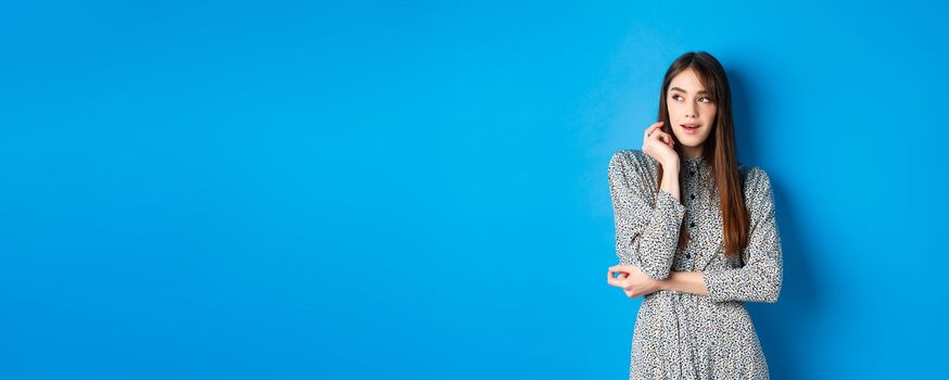 Pensive caucasian girl with long natural hair, wearing vintage dress, looking left with thoughtful face, standing on blue background.