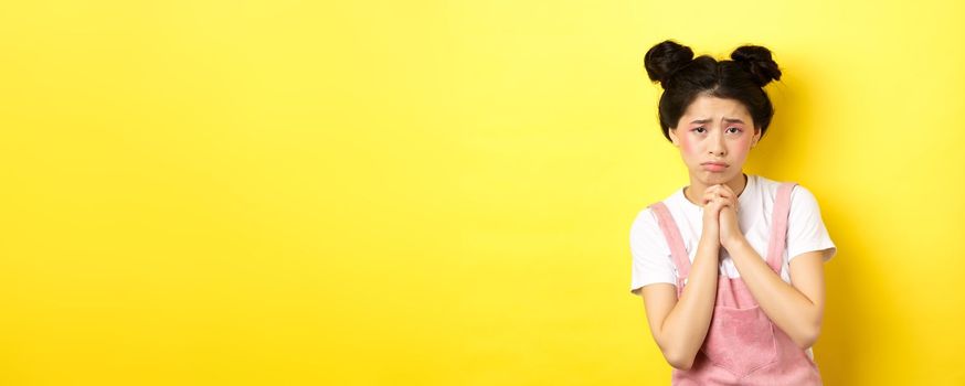 Sad asian girl pleading for help, begging you, sulking and whining, standing on yellow background.