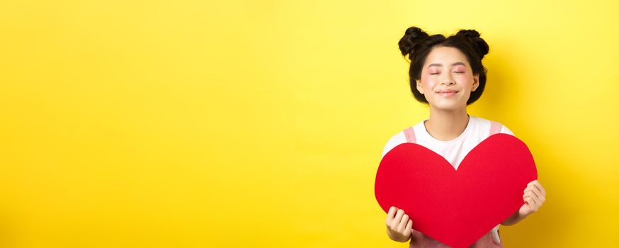 Cute teenage asian girl dreaming of true love, close eyes and showing big red heart cutout, waiting for soulmate on Valentines day, standing with romantic makeup on yellow background.