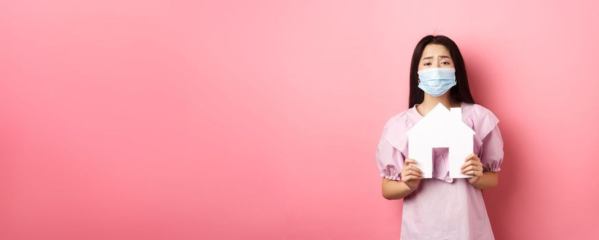 Real estate and pandemic concept. Begging asian girl showing paper house cutout, wearing medical mask, asking for apartment, standing against pink background.