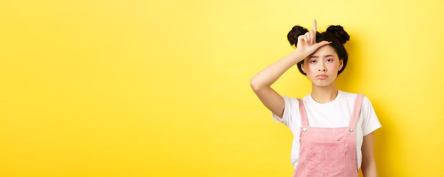 Sad girl showing loser sign on forehead and sulking upset, feeling disappointed in herself, standing on yellow background.