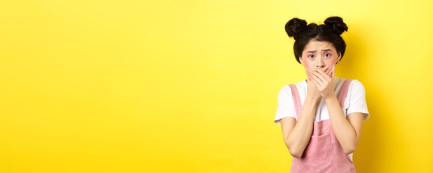 Worried asian teen girl covering mouth with hands, looking concerned and anxious, standing scared on yellow background with glam makeup and summer clothes.