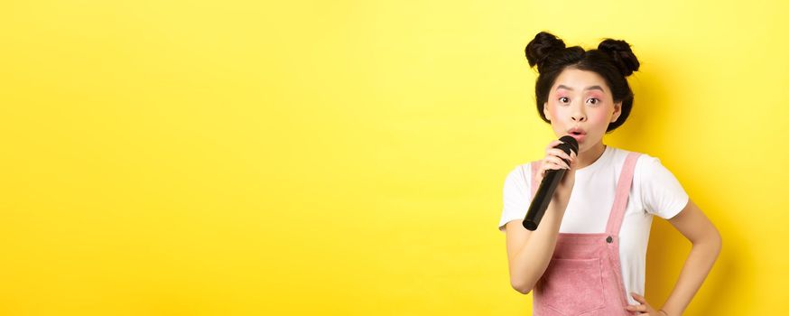 Cute asian teen girl with bright makeup, singing in microphone karaoke, standing against yellow background.