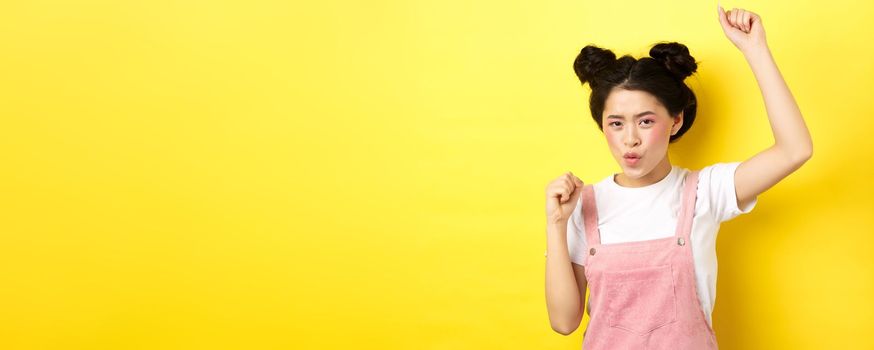 Excited asian woman getting motivation, raising hand up and chanting, celebrating victory, triumphing and standing on yellow background.