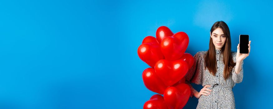 Valentines day. Beautiful young woman with makeup, wearing trendy dress, standing near red hearts balloons and showing empty mobile screen, blue background.