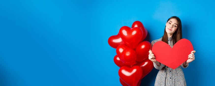 Valentines day. Romantic girl in dress showing big red heart cutout, dreaming of love, standing near holiday balloons on blue background.