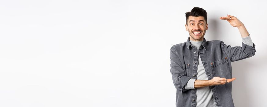 Cheerful guy smiling and showing big size, showing large thing and looking excited, standing against white background.