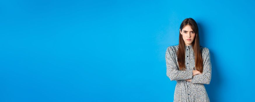 Angry frowning woman in dress, cross arms on chest and feel offended about something unfair, standing upset against blue background.