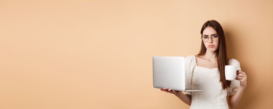 Tired and reluctant girl hates working, drinking morning coffee and using laptop, frowning upset, standing on beige background.